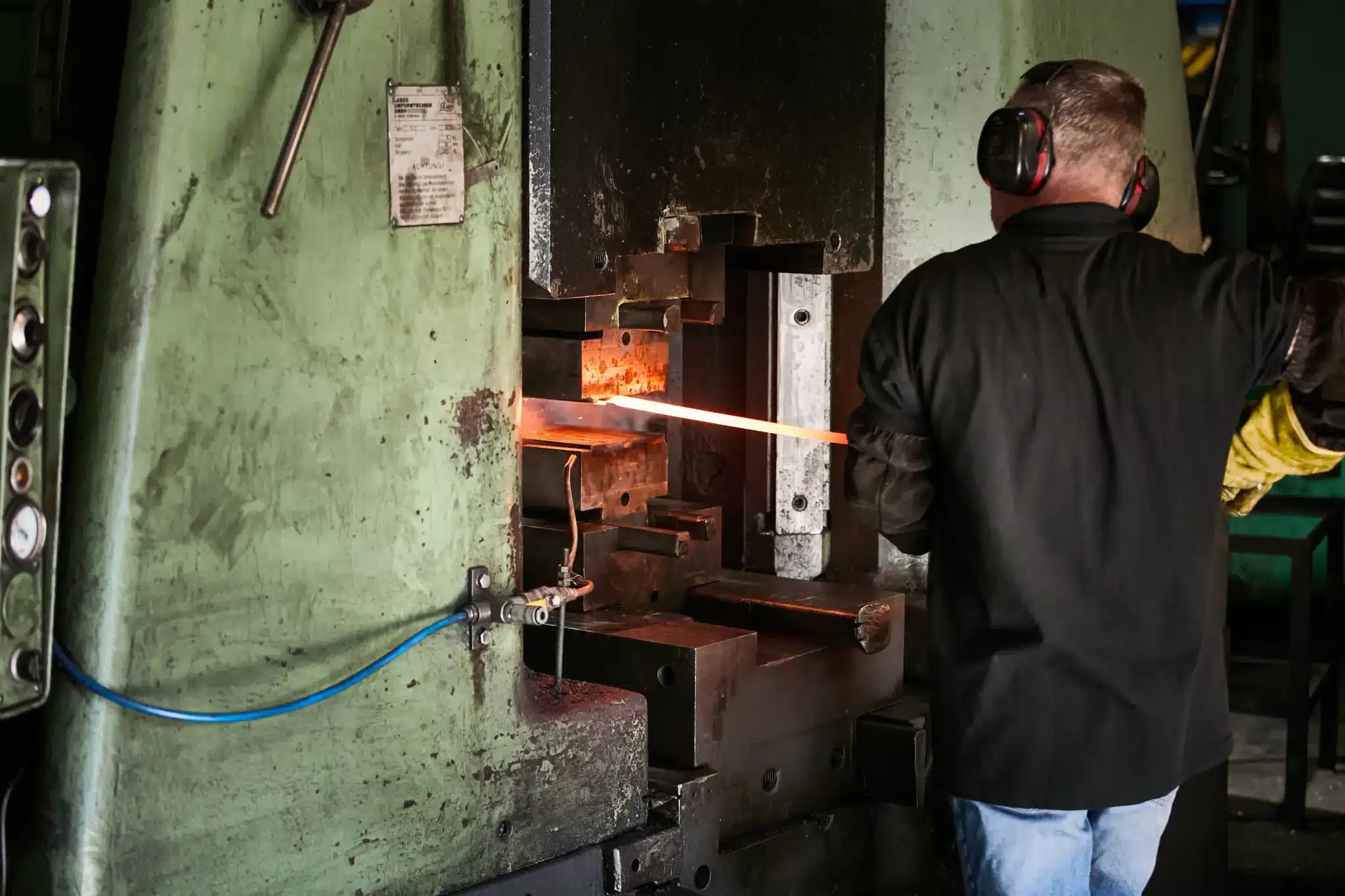 A forger in ear defenders feeding a glowing orange-hot steel bar into the dies of the LASCO press to hot drop forge an Ernest Wright Nurseryman Scissor blade in Sheffield.