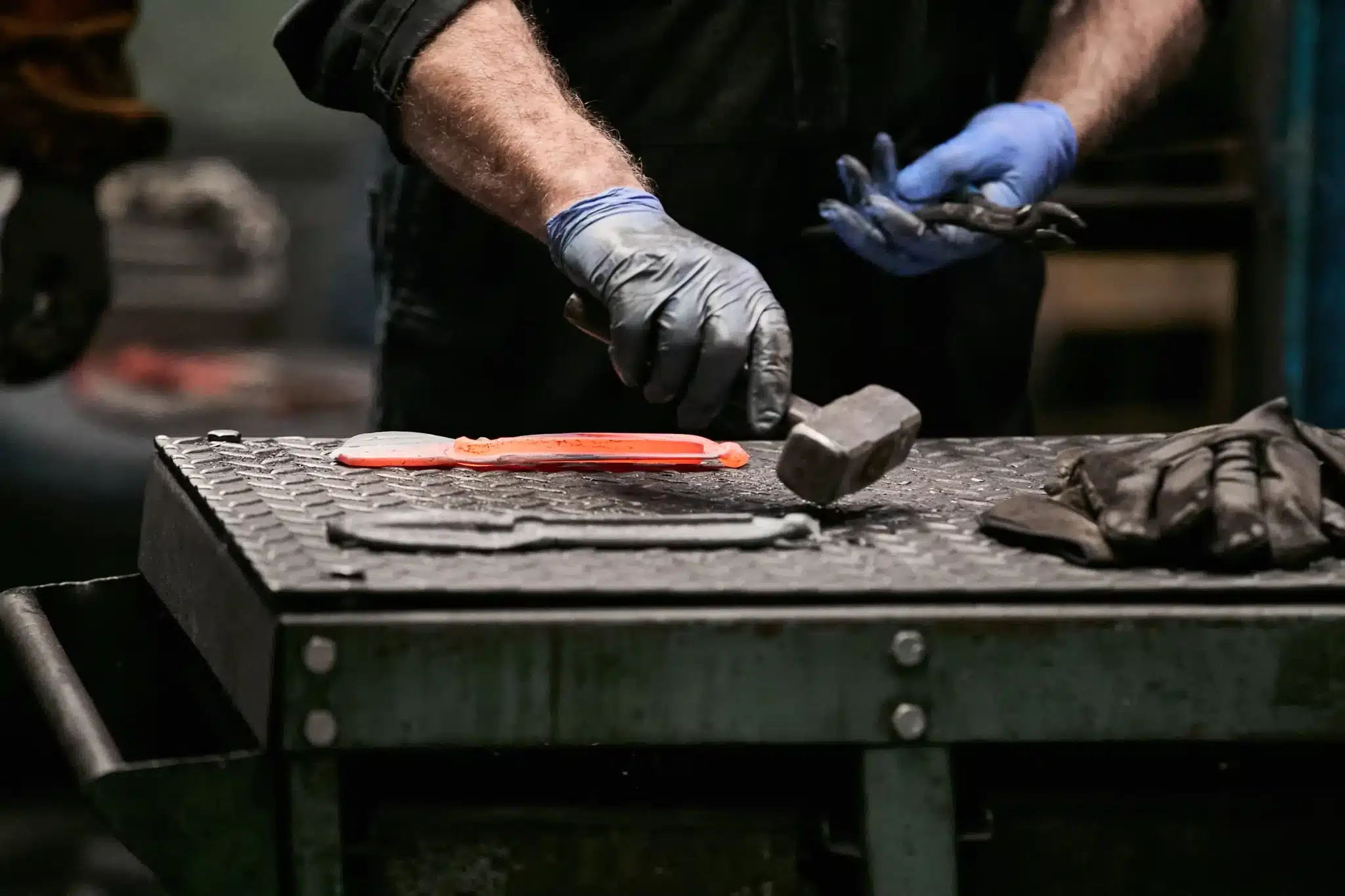 A glowing orange hot-forged Nurseryman Scissor blank resting on the bench beside a hammer, still bearing the flash of excess steel pressed out by the forging dies.