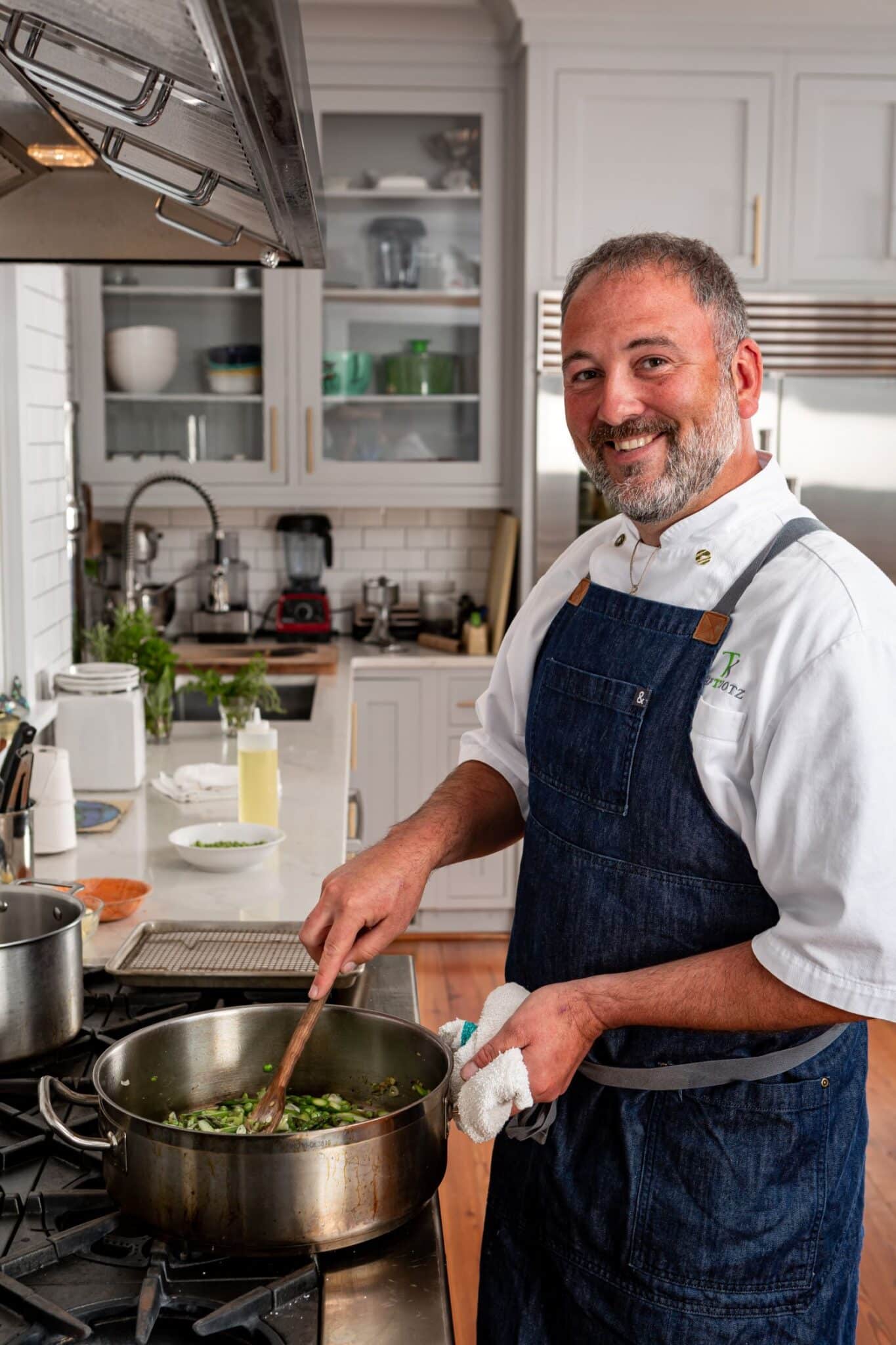 Private chef Ty Kotz in chef's whites and a denim apron, stirring a pan on the stove of a home kitchen