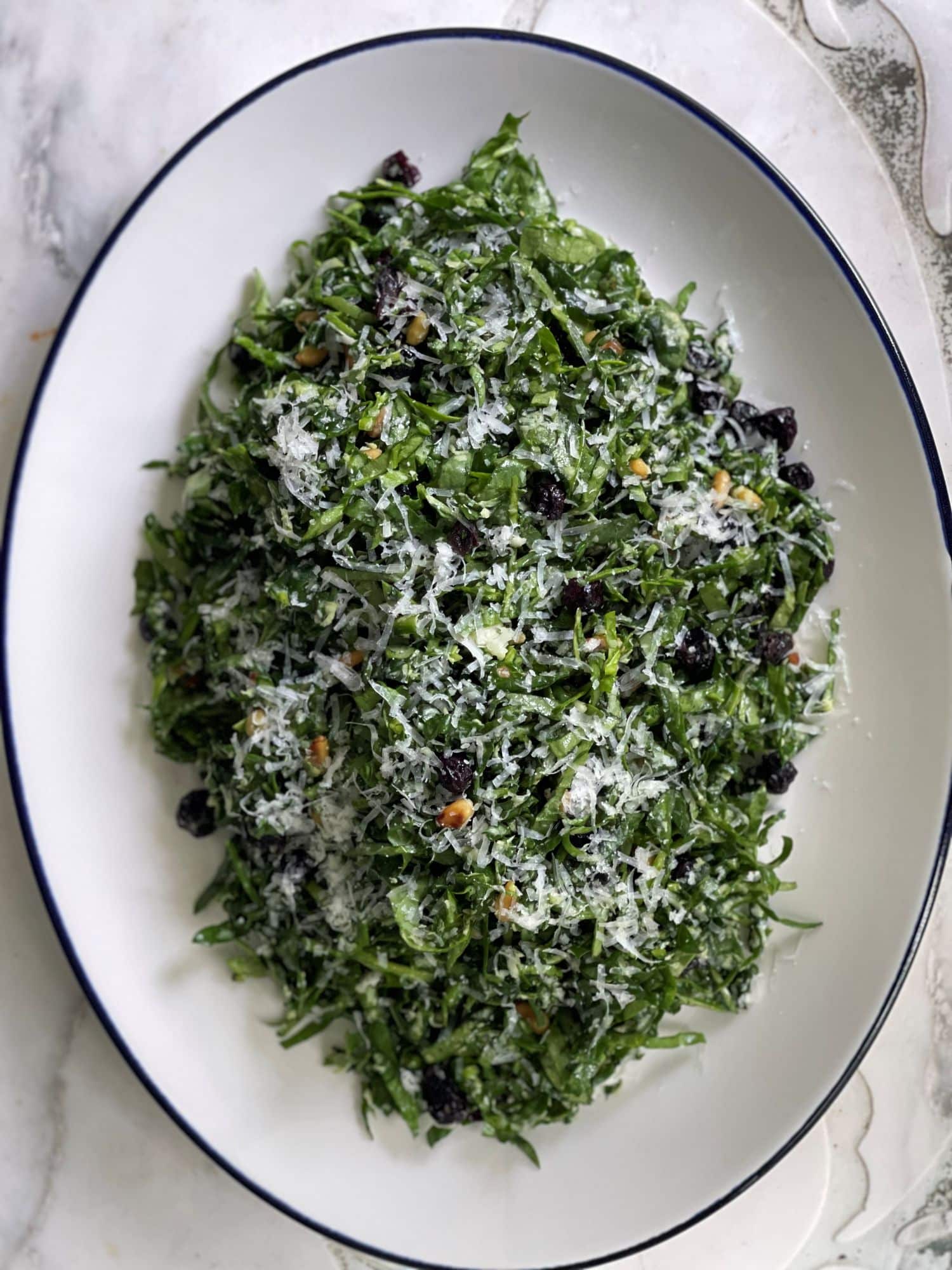 A white oval platter of finely shredded greens dressed with grated cheese and dark dried fruit