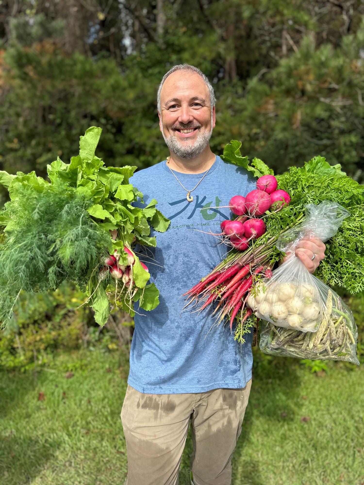 Ty Kotz outdoors in a garden, holding armfuls of freshly picked radishes, carrots, dill and leafy greens