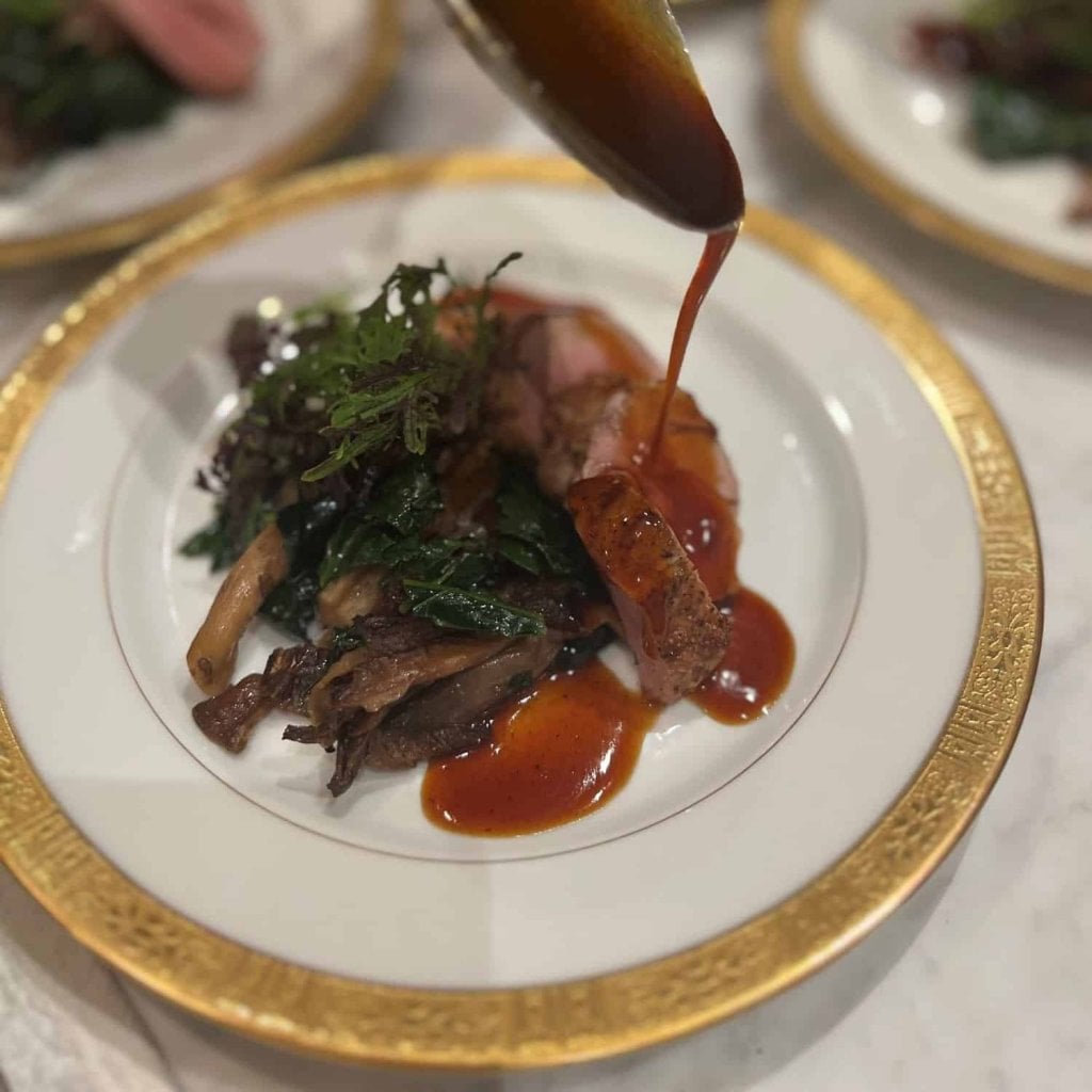 A glossy sauce being poured over sliced meat, mushrooms and wilted greens on a gold-rimmed plate