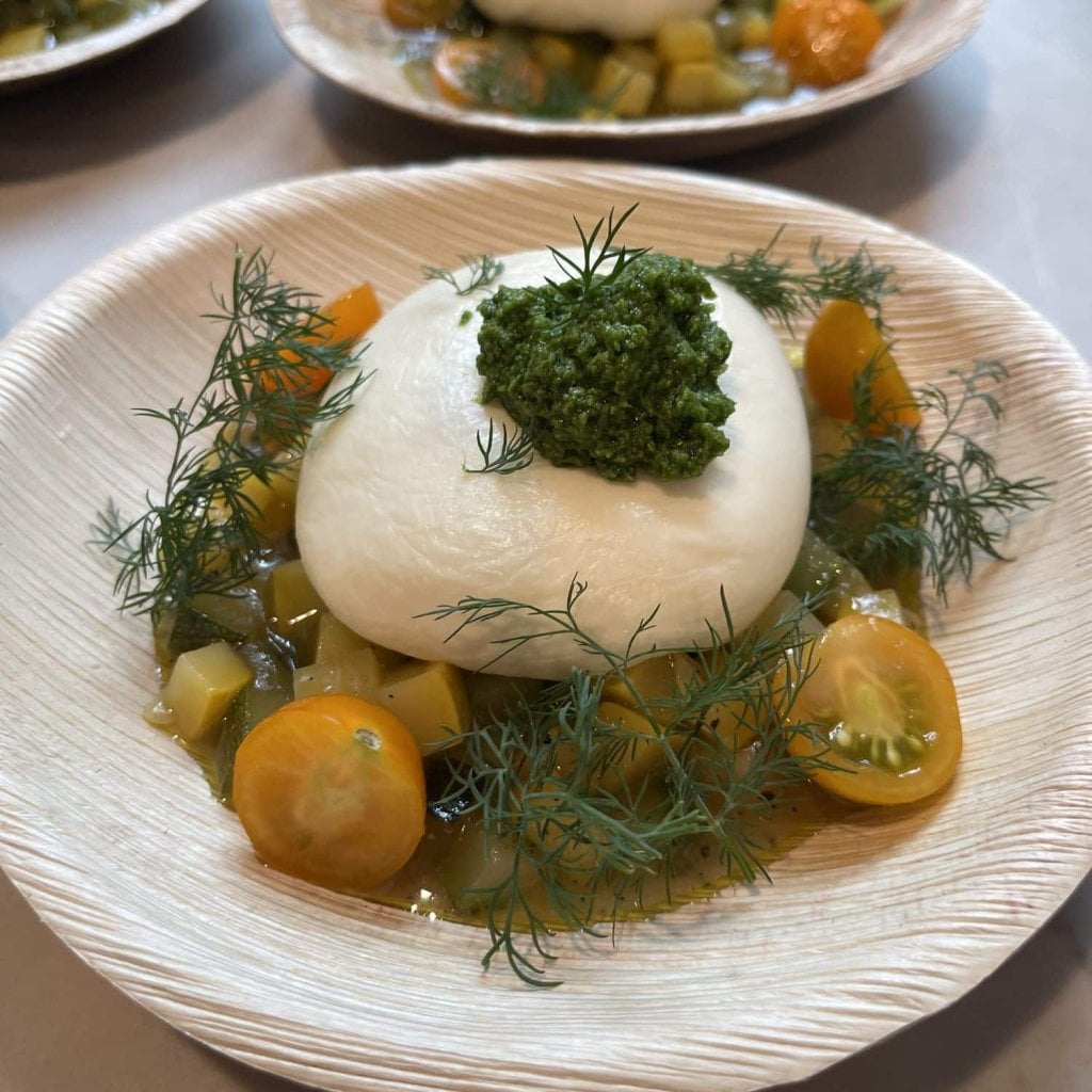 A ball of burrata topped with bright green herb sauce, in a light broth with diced squash, halved yellow cherry tomatoes and sprigs of dill