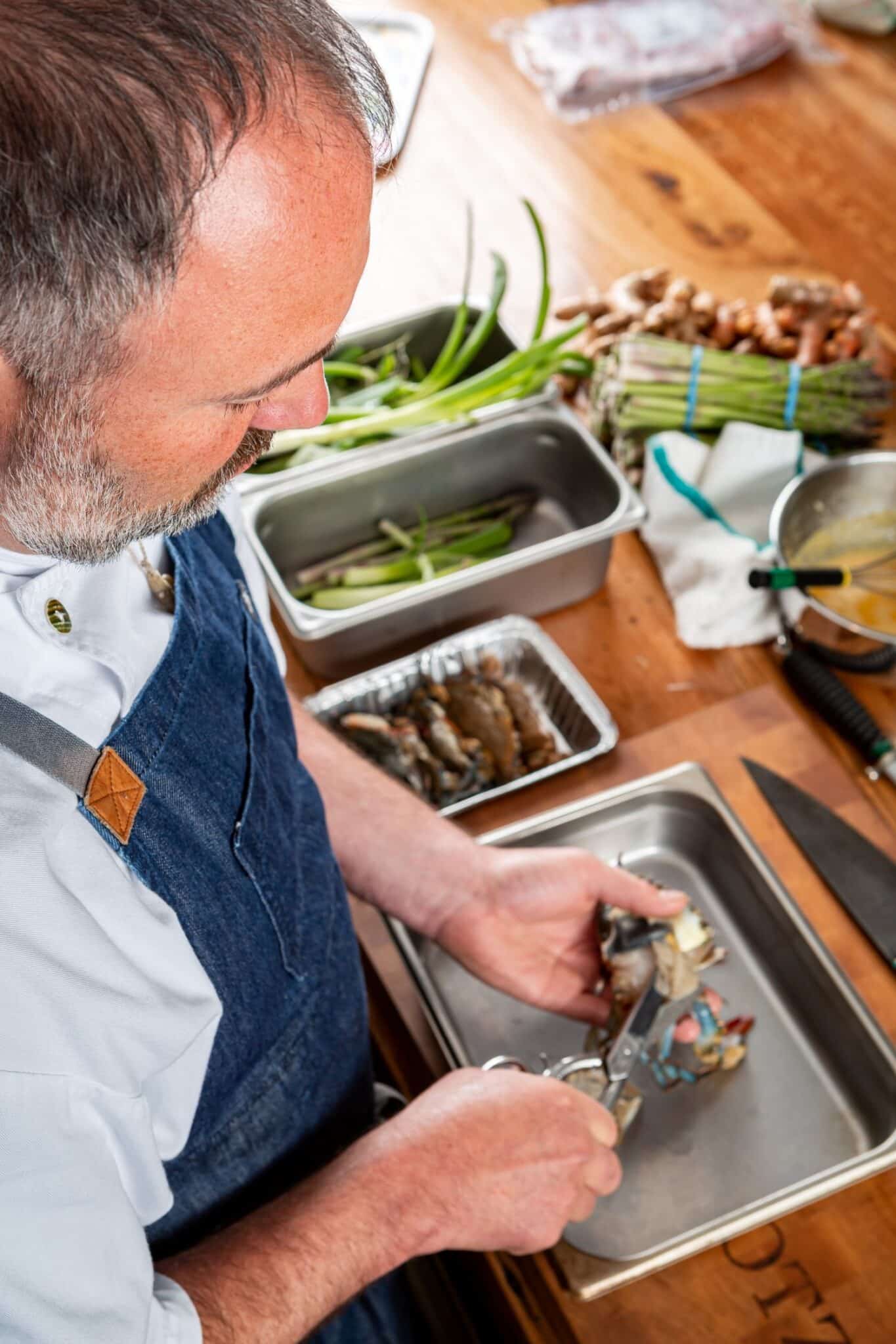 Ty Kotz cutting a blue crab with kitchen scissors over a steel tray, beside trays of spring onions, asparagus and more crab